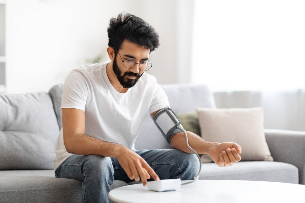A young adult male with dark hair and facial hair sits on a couch to check his blood pressure. He wears glasses and is dressed in a white tshirt and jeans. 